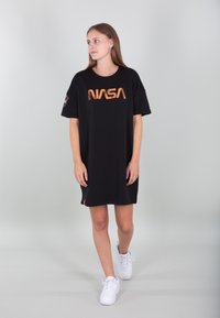 Crna oversized majica haljina s narančastim NASA logotipom, kratkim rukavima i bočnim akcentom logotipa. Nosena s bijelim tenisicama na svijetloj pozadini.