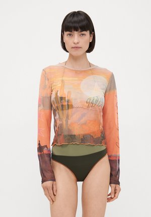 Langermet mesh topp med et ørkenlandskap trykk i oransje, med en rund hals og volang nederkant. Brukt over olivengrønne høyt midje bukser.