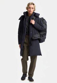 Lungo cappotto imbottito nero con cappuccio, indossato sopra una maglia nera e pantaloni verde oliva. Grande zaino nero con dettagli in rete. Scarpe da ginnastica nere.