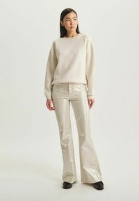 Sweatshirt beige clair à coupe décontractée, associé à un pantalon évasé crème taille haute brillant avec trois boutons sur le devant. Chaussures noires.
