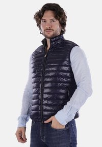 Gilet impermeabile blu navy con colletto alto, design trapuntato, chiusura con zip e tasche laterali, indossato sopra una camicia a maniche lunghe azzurra.