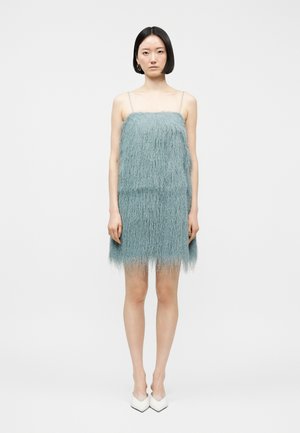 Femeie în picioare, purtând o rochie texturată fără mâneci de culoare albastru deschis, cu bretele subțiri și pantofi cu toc alb, cu vârf ascuțit, pe un fundal alb simplu.