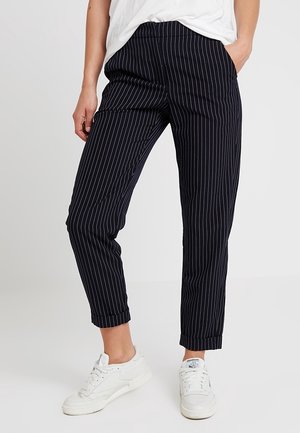 Persona con pantalones cortos negros con rayas finas y zapatillas blancas, manos en los bolsillos, sobre un fondo claro y liso.