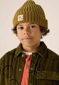 Olijfgroene geribbelde breimuts met een patch, gecombineerd met een corduroyjas met cirkelpatronen en een rode overhemd eronder.