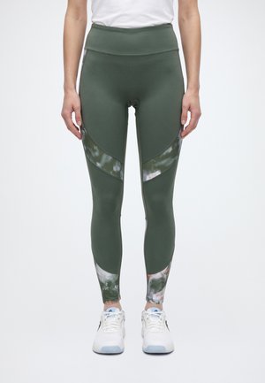 Un modèle portant un legging vert foncé taille haute à empiècements à motifs et des chaussures de sport blanches se tient devant un fond uni.