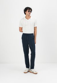Weißes Poloshirt mit kurzen Ärmeln und strukturiertem Muster, kombiniert mit navy Jogginghosen und cremefarbenen Sneakers mit schwarzen Akzenten. Das Model trägt dunkle Sonnenbrillen.
