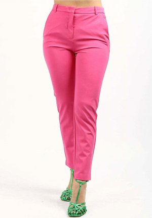 Kobieta w jasnoróżowych, dopasowanych spodniach i metalicznie zielonych sandałach na wysokim obcasie z paskami stojąca na białym tle.