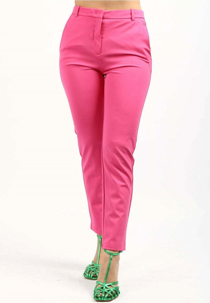 Femme portant un pantalon rose vif ajusté et des sandales à talons hauts métalliques vertes à brides, debout devant un fond blanc.
