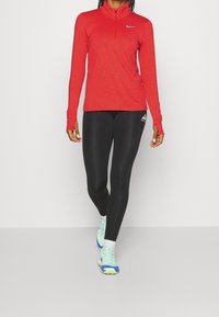 Rood zip-up top met lange mouwen en een Nike-logo, gecombineerd met zwarte leggings met een wit driehoeklogo, en hardloopschoenen met blauwe accenten.