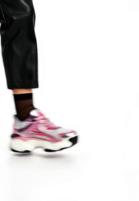 Persona con pantalones negros recortados, calcetines negros transparentes y zapatillas gruesas blancas, rosas y moradas caminando sobre un fondo blanco.