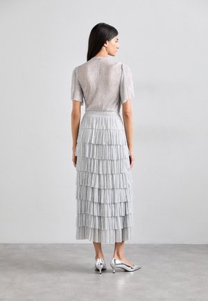 Vestido plateado con mangas cortas abullonadas, que presenta una falda en capas. Combinado con elegantes zapatos de tacón plateados.