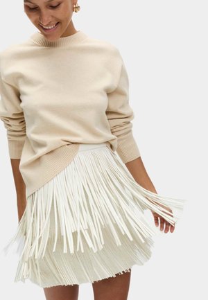 Beige gebreide trui met geribde mouwen, gecombineerd met een witte rok met franjes met verticale strepen. De outfit heeft een gelaagd ontwerp.