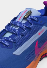 Μπλε και πορτοκαλί παπούτσι για τρέξιμο σε μονοπάτι με μπλε κορδόνια με σχέδια, ετικέτα αδιάβροχου Gore-Tex και κείμενο "Pegasus Trail" στη γλώσσα.