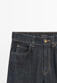 Jeans in denim scuro con taglio dritto, dotati di una cintura, tasca frontale e bottoni in oro. Dettagli di cuciture a contrasto.