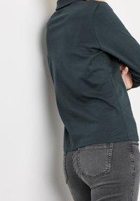 Mörkgrön långärmad polotröja med krage. Tyget ser glatt ut med minimala sömmar, i kombination med åtsittande grå jeans med bakfickor.