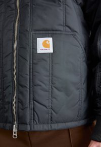 Czarna pikowana kurtka z srebrnym zamkiem błyskawicznym i pomarańczową naszywką z logo. Posiada boczną kieszeń i ściągacze przy mankietach dla dodatkowej tekstury.