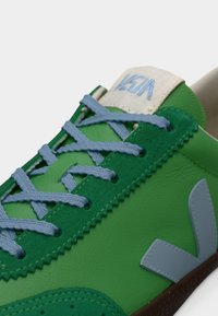 Baskets vertes avec une tige en cuir texturé, des lacets bleu clair et un logo gris. Elles disposent d'une semelle en caoutchouc et de détails cousus.