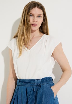 Jeune femme aux longs cheveux blonds portant un haut blanc sans manches et un pantalon en jean bleu taille haute avec une ceinture nouée, debout contre un mur blanc.