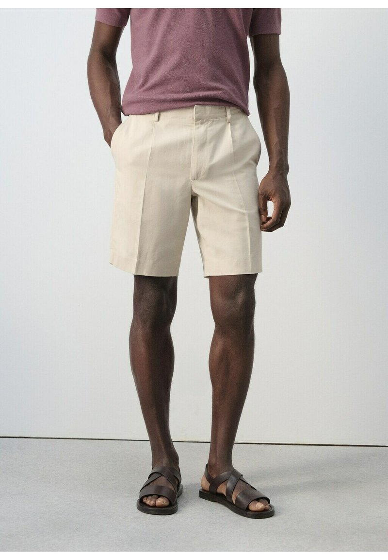 Homme portant un short beige mi-cuisses, une chemise mauve à manches courtes et des sandales marron foncé à lanières croisées, debout les mains dans les poches.