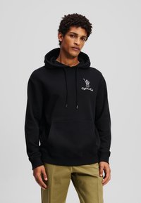 Schwarze Baumwollkapuzenjacke mit einer frontalen Tasche, Kordelzugkapuze und weißem Grafikdetail auf der linken Brust. Kombiniert mit kaki Farben Hosen.