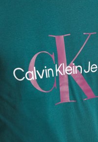Tyrkysové bavlněné tričko s velkým růžovým nápisem "Calvin Klein Jeans". Materiál má hladkou texturu a přiléhavý střih.