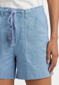 Modré lněné kraťasy se stahovacími šňůrkami v pase, dvěma předními kapsami a měkkou texturou. Obsahují třásně jako akcenty na stahovací šňůrce.