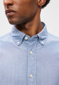 Πουκάμισο με κουμπιά σε ανοιχτό μπλε χρώμα με λευκές κατακόρυφες ρίγες, διαθέτοντας γιακά που κουμπώνει και δύο κουμπιά στη μπροστινή σέλα.