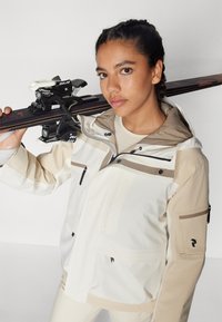 Skijakke i hvid og beige, med flere lynlåslommer, justerbar hætte og velcro-manchetter. Inkluderer et ski i hånden med bindinger.