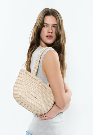 Joven mujer con cabello largo y castaño, vistiendo una camiseta gris sin mangas, sosteniendo un bolso tejido beige en el hombro sobre un fondo blanco.
