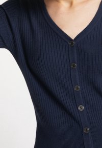 Granatowy kardigan z prążkowanego dzianiny, z dekoltem w kształcie litery V, długimi rękawami i z przodu zapinany na guziki. Gładka tekstura, dopasowany krój i delikatny stretch.