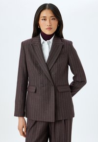 Tumma ruskea pinstripe-bleiseri, jossa on kaksirivinen nappikiinnitys, olkatoppaus, läpälliset taskut sekä silkkinen kangas, jossa on kontrastiväriset ohuet raidat.