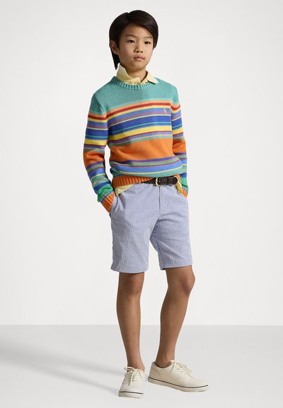 Pulover multi-color cu dungi în turcoaz, portocaliu, galben și albastru, purtat peste o cămașă galben deschis. Asortat cu pantaloni scurți cu dungi albastre și albe și pantofi bej.