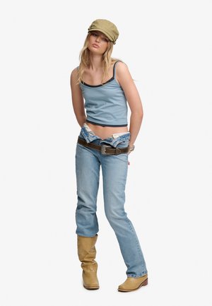 Ung kvinde iført en kaki kasket, lyseblå tanktop, lavtaljede jeans med brunt bælte og tanfarvede støvler, står med hænderne bag ryggen.