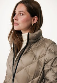 Gevoerde, beige pufferjack met hoge kraag, zwarte rits en glanzende textuur, gedragen over een witte coltrui met diamanten stiksel.
