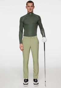 Långärmad grönmönstrad golfskjorta med hög krage, i kombination med ljusgröna byxor, svartvita golfsko och en golfklubba.