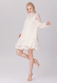 Robe en dentelle blanche avec un motif floral texturé, longues manches transparentes, ourlet volanté et bords festonnés, portée avec des sandales à talons blanches.