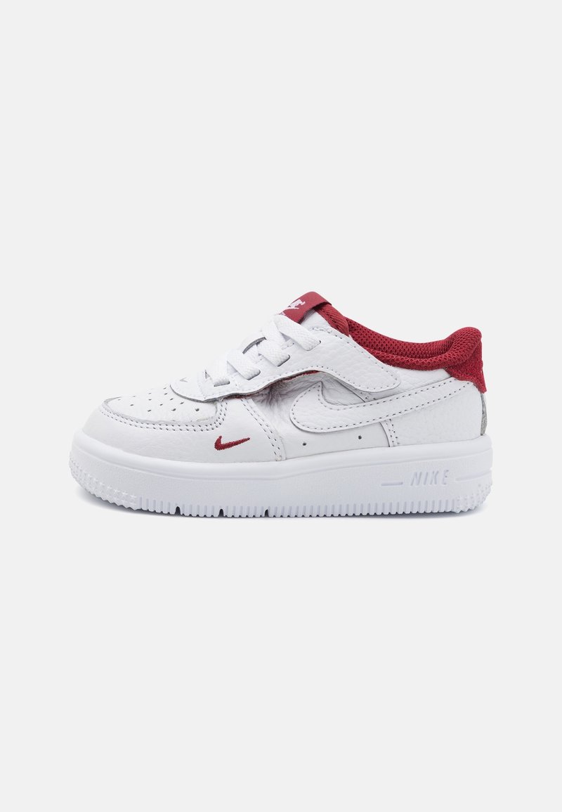 Baskets basses blanches Nike avec doublure intérieure rouge, accents du logo swoosh rouge, lacets et une bride velcro sur un fond blanc uni.