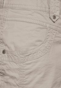 Vaaleanbeige puuvillakangas, jossa on sivutasku, jossa on erottuva tikkaus ja pyöreä metallinen napituskoriste. Sileä rakenne.