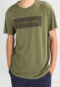 Camiseta de algodón verde oliva con un gráfico rectangular oscuro que presenta el texto "G-STARD RAW", mangas cortas y un corte relajado.