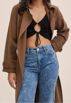 Femme portant un manteau marron, un crop top noir avec une découpe à l'avant et un nœud, ainsi qu'un jean bleu taille haute avec un motif paisley subtil.