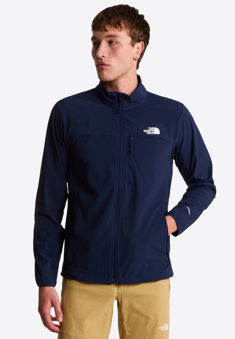 Chaqueta softshell azul marino con cuello alto, bolsillos con cremallera y detalles del logo en blanco. Textura suave con un diseño entallado.