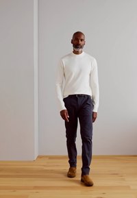 Balta ilga rankovėmegztinis, tamsios kelnės ir rudi zomšiniai bateliai. Stovi ant medinių grindų, su šviesiu fonu. Paprastas, modernus dizainas.