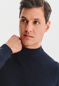 Granatowy golf wykonany z dzianiny. Dopasowany fason z prążkowanym kołnierzem. Osoba na zdjęciu ma krótko uczesane włosy i neutralny wyraz twarzy.