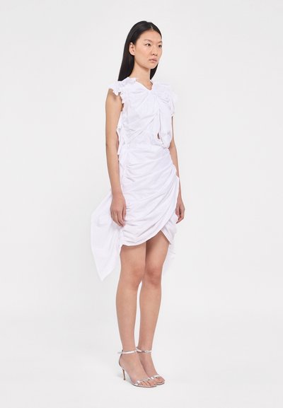 Robe blanche avec des épaules à volants, un corsage tordu et des détails froncés. Le tissu lisse drape de manière asymétrique ; associé à des sandales à talons transparents.