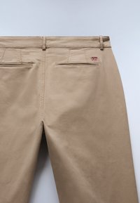Beiga byxor med en slät textur, som har två bakfickor, subtila accentstygn och en liten norsk flagga nära höger ficka.