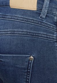 Jeans de denim azul-escuro com uma textura suave, apresentando costuras decorativas e um pequeno bolso frontal com um botão de pressão prateado.