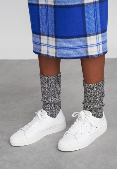 Valkoiset tennarit, joissa on teksturoitu muotoilu ja nauha-accentit, paritettu harmaiden ribbimeikkisukkien kanssa. Sininen ruudullinen hame näkyy päältä.