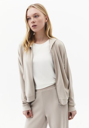 Blonde vrouw die een beige hoodie met rits en bijpassende broek draagt over een wit shirt, staand tegen een effen witte achtergrond.