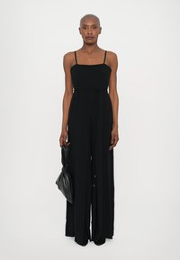 Black, sleeveless jumpsuit with a square neckline, wide leg design, and a tie detail at the waist. Accompanied by a sleek black bag.