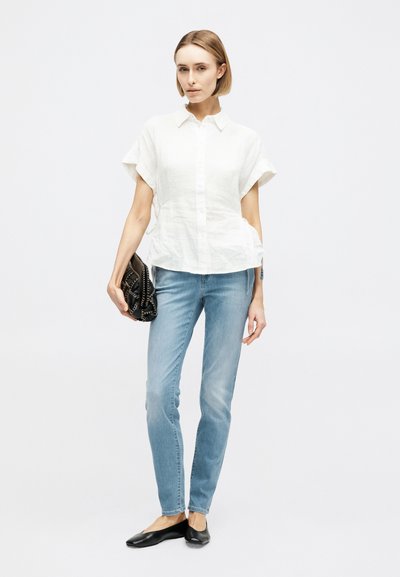 Femme portant une chemise blanche à manches courtes et boutons, un jean bleu clair, des ballerines noires, et tenant un sac à main noir avec des détails en chaîne.